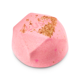 Kochi. A light pink, half bath bomb with a flat base in a geometric dome shape. The top is sprinkled with textured brown granules and features embossed "LUSH" text, with a layer of pink at the base.