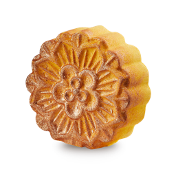 Moon Cake bath bomb. A golden yellow bath bomb with scalloped edges and a detailed, embossed floral pattern on its top. It is coated in shimmering gold lustre.