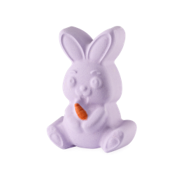 The Munching Bunny bath bomb is a seated lavender-purple rabbit with large ears, holding a bright orange carrot. It features a cute, carved face and is set against a clean, neutral grey background.
