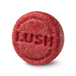 New shampoo. A circular, highly textured red solid shampoo bar. The fibrous, shredded surface features the brand name 'LUSH' embossed clearly in the centre of the round face.