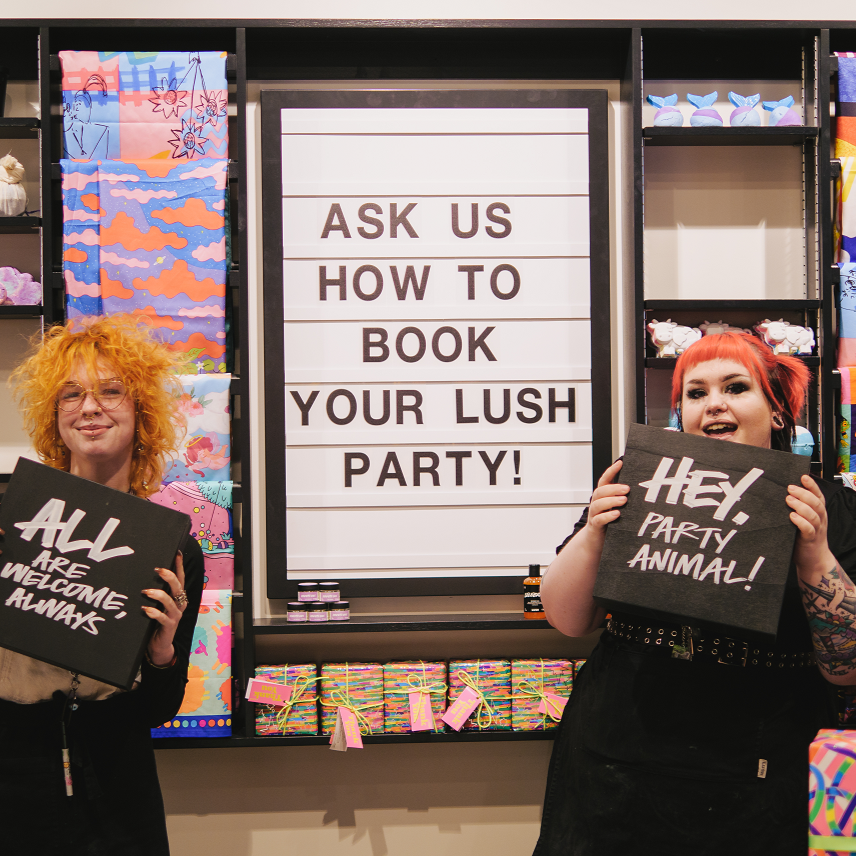 Two members of staff dressed in black holding signs that read All Are Welcome Always, and Hey Party Animal. They are stood either side of a cinema style display sign that reads Ask Us How To Book Your Lush Party!
