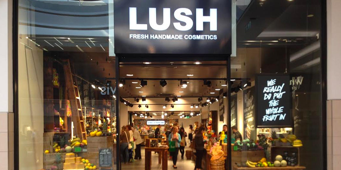 Newcastle shop front. Exterior view of shop. Black framed shop with glass frontage. Large entrance in centre with illuminated Lush sign above. Products displayed in windows either side of entrance.