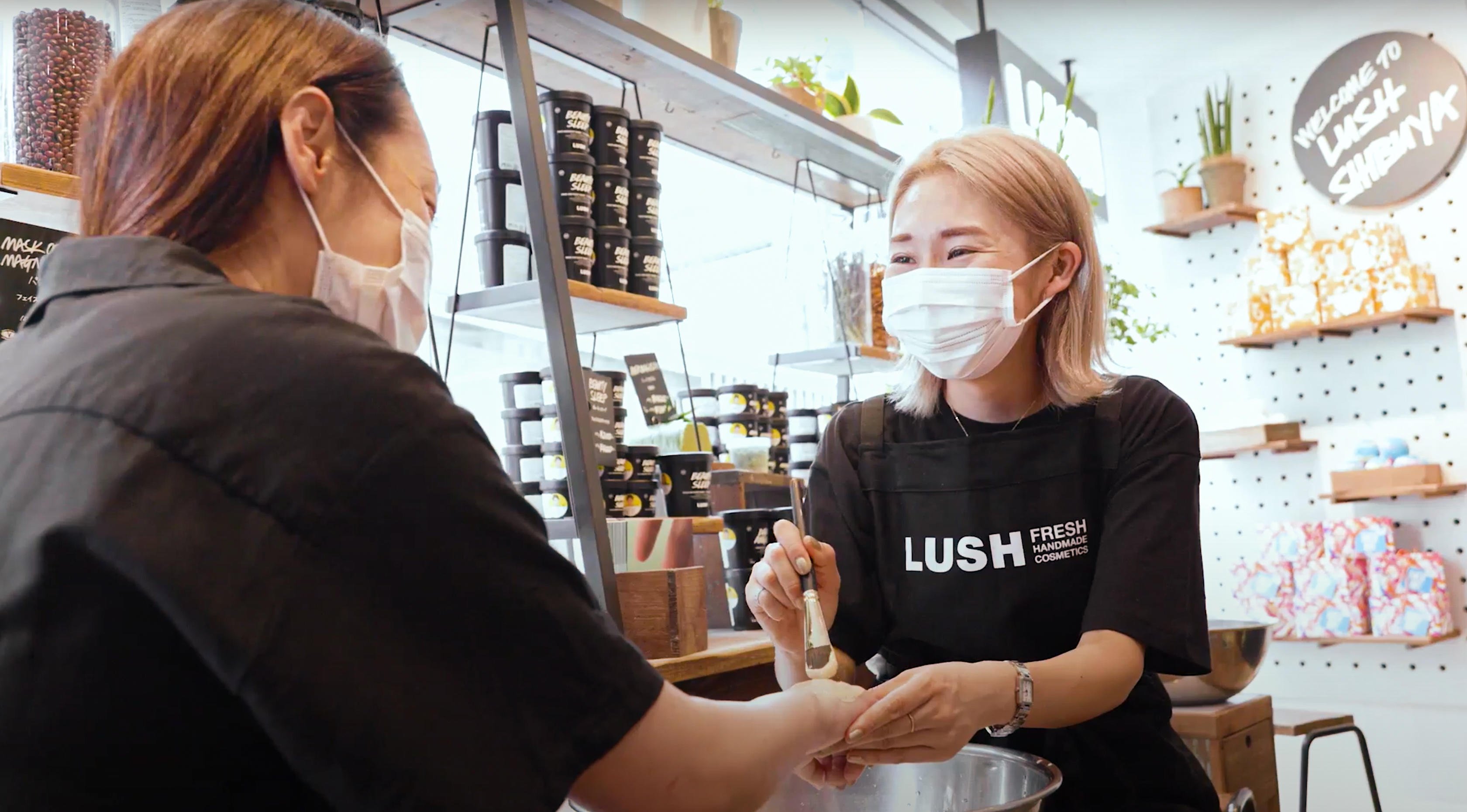 Member of staff wearing a facemask applying product with a brush to a customers arm. Displays of various packaged and unpackaged products in the background.