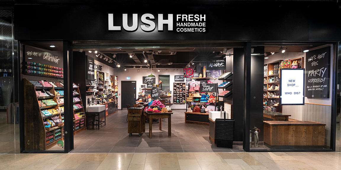 Peterborough Store Front. Exterior view of shop. Black framed shop with glass frontage. Large entrance in centre with illuminated Lush sign above. Full interior of the shop is visible from the outside.