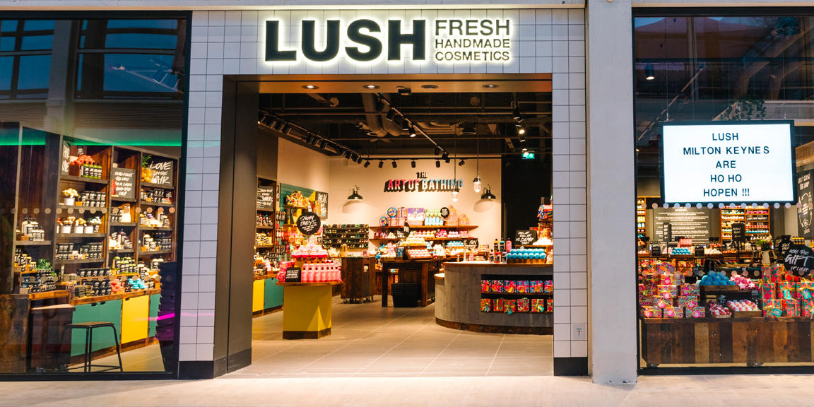 Milton Keynes shop front. Exterior view of shop. Tile framed shop with glass frontage. Large door way in centre with illuminated Lush sign above. Products displayed in windows either side of entrance.