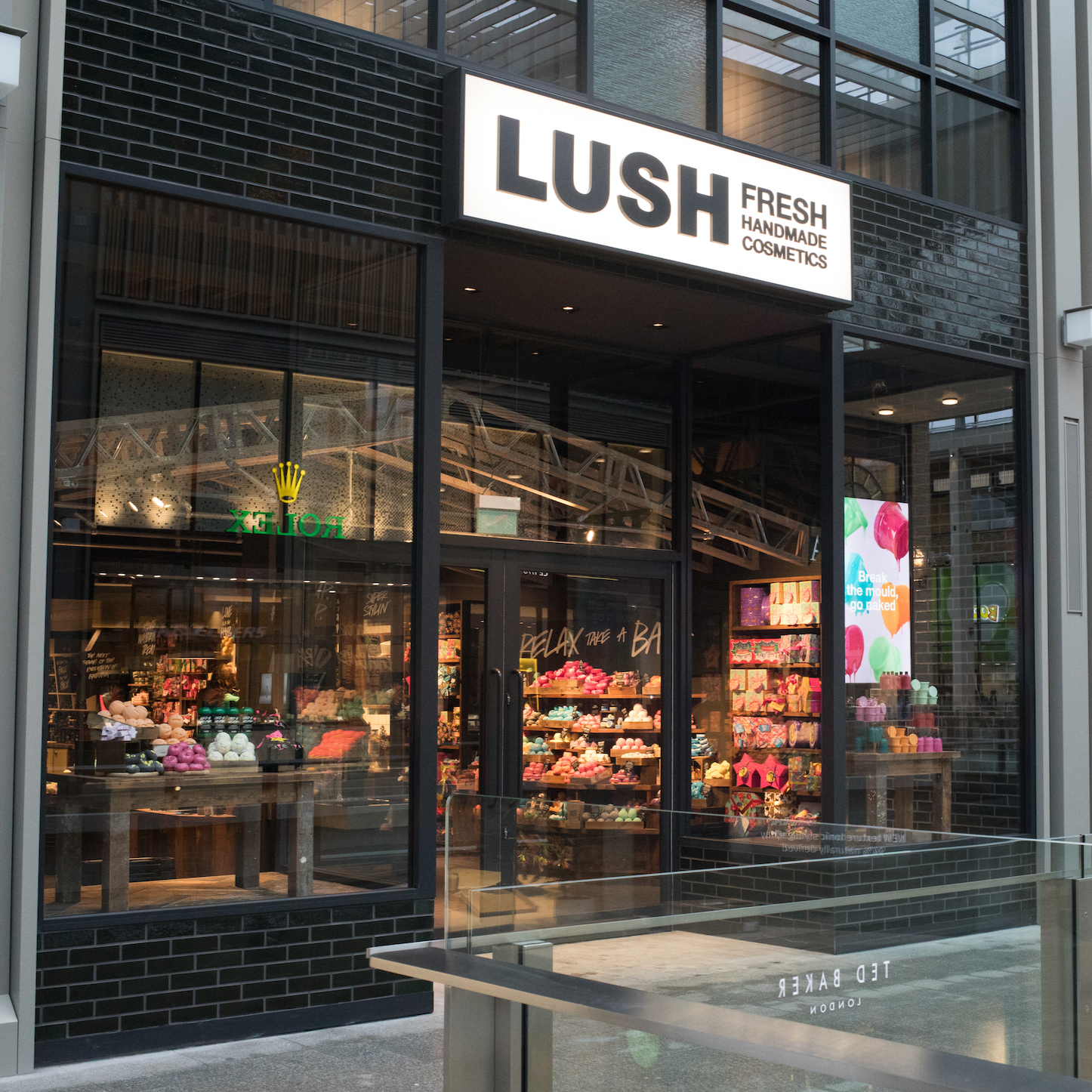 Oxford Shop Front. Exterior view of shop. Black framed shop with glass frontage. Large entrance in centre with illuminated Lush sign above. Products displayed in windows either side of entrance. Large  illuminated lightbox in window to right of entrance.