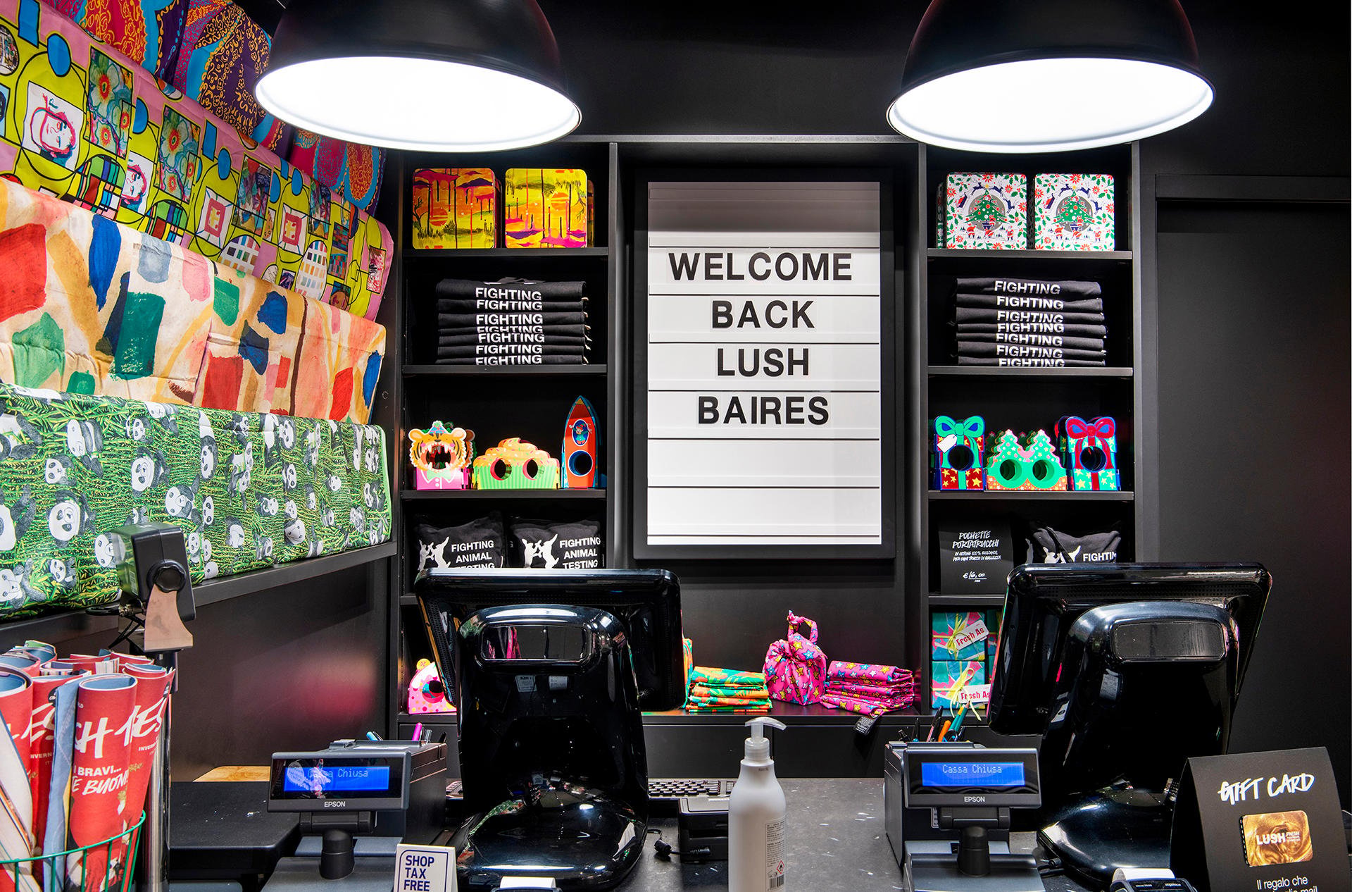 Interior view of shop. Tillpoint in foreground. Products displayed on wall behind. Large cinema style illuminated display reads Welcome Back Lush BAires. Knotwraps displayed on wall to the left.