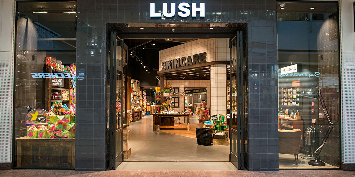 The front of the store at Cribbs Causeway. There are two columns either side of open double doors, with a large lit up display saying LUSH. The shop looks warm and inviting.