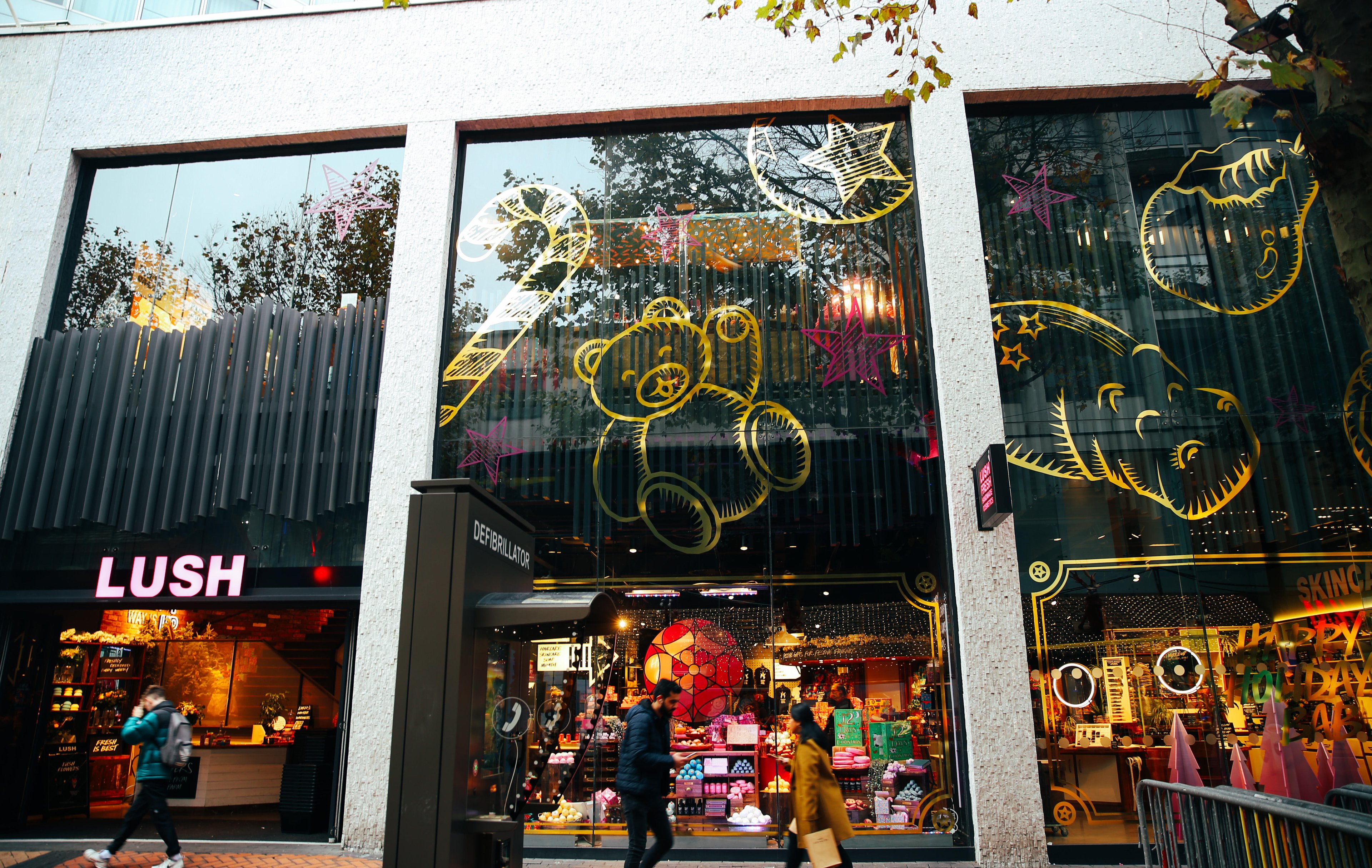 Exterior view of shop. Black and white framed shop with glass frontage. Large entrance on left with illuminated Lush sign above. Products displayed in windows running along length of shop.