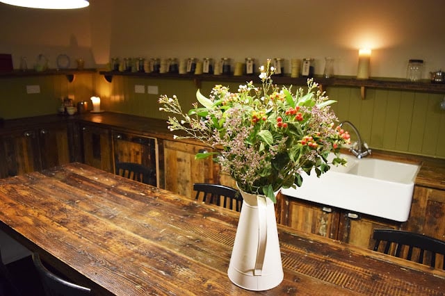 Interior view of the Spa. In the foreground is a white Jug with a bouquet of fresh flowers sat on a wooden table. In the background along the wall is a shelf containing various glass jars with individual hand written tags.