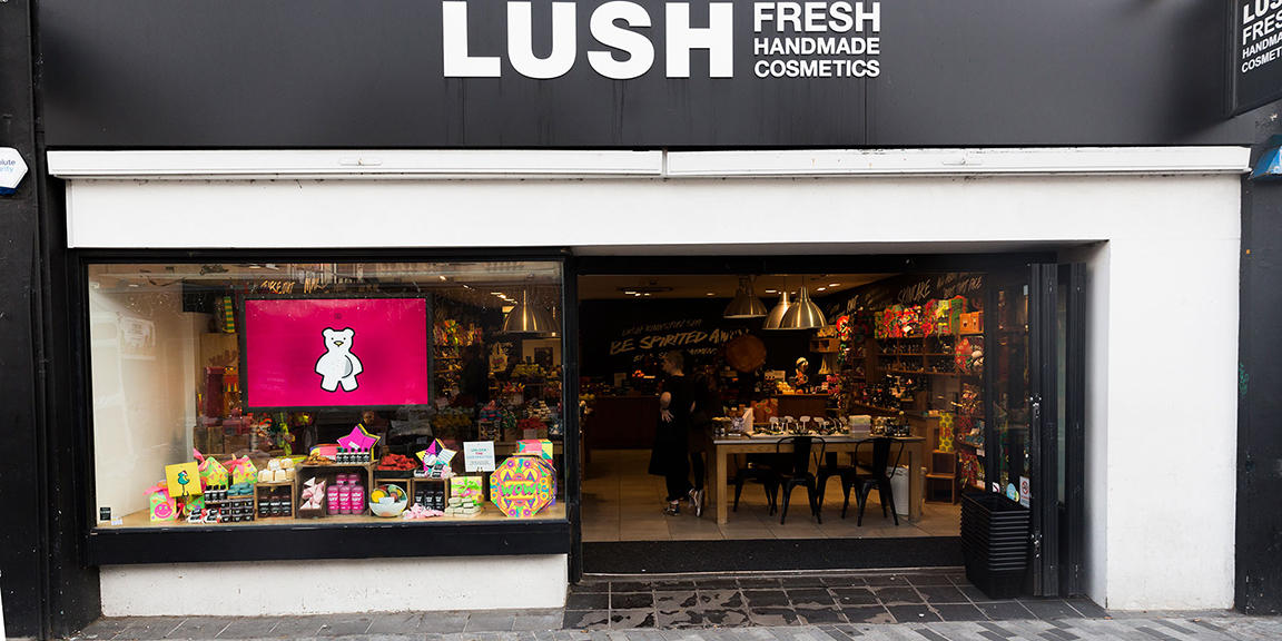 Kingston shop front. Exterior view of shop. Black and white framed shop with glass frontage. Large door way on right with Lush sign above. Products displayed in windows on right of entrance.