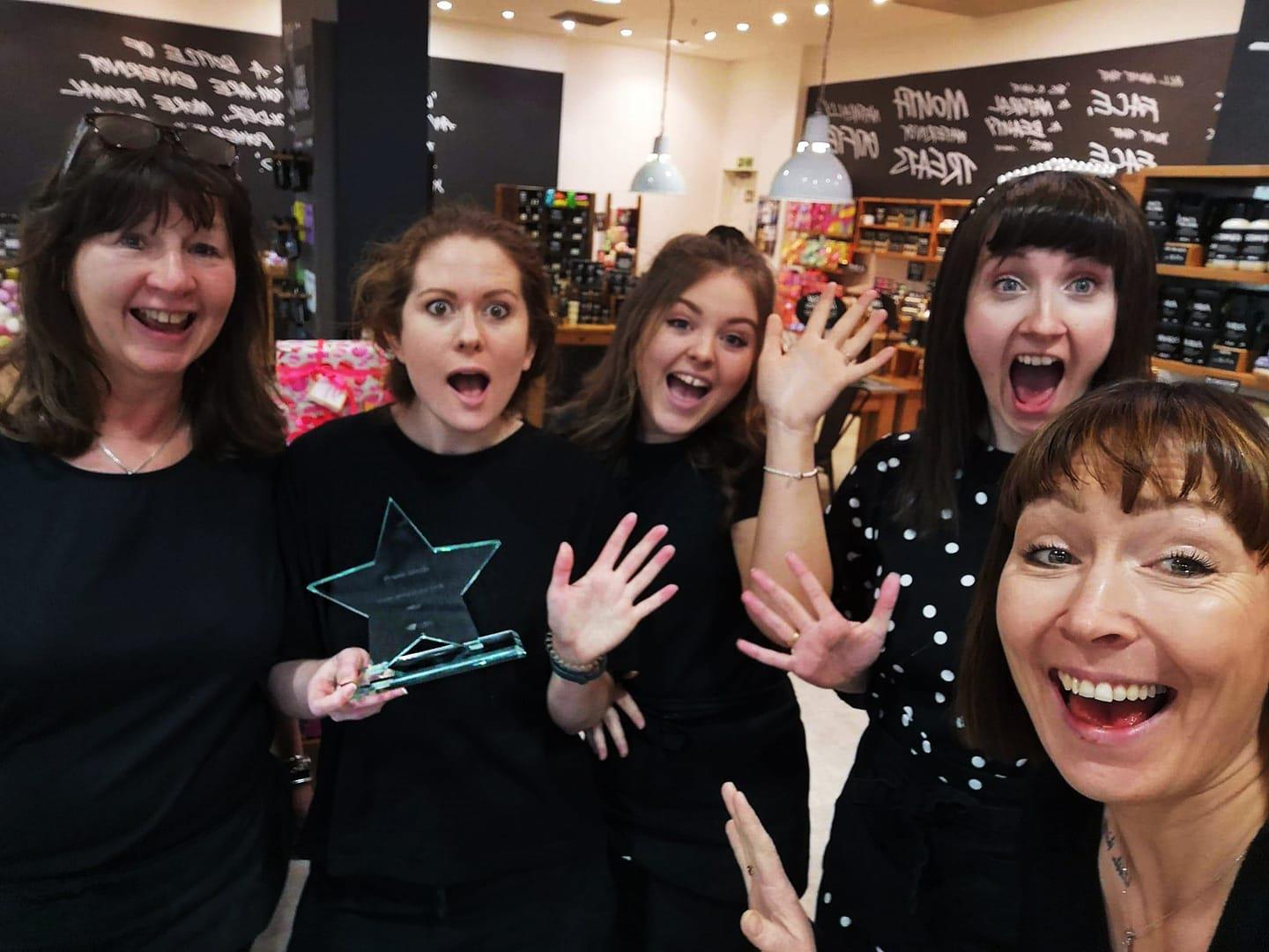 5 members of staff dressed in black smiling and waving at the camera. Shop displays visible behind.