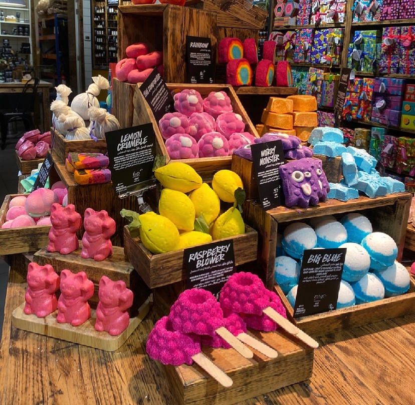 Various differently coloured and shaped unpackaged bath products sat in wooden pens and on wooden display units.