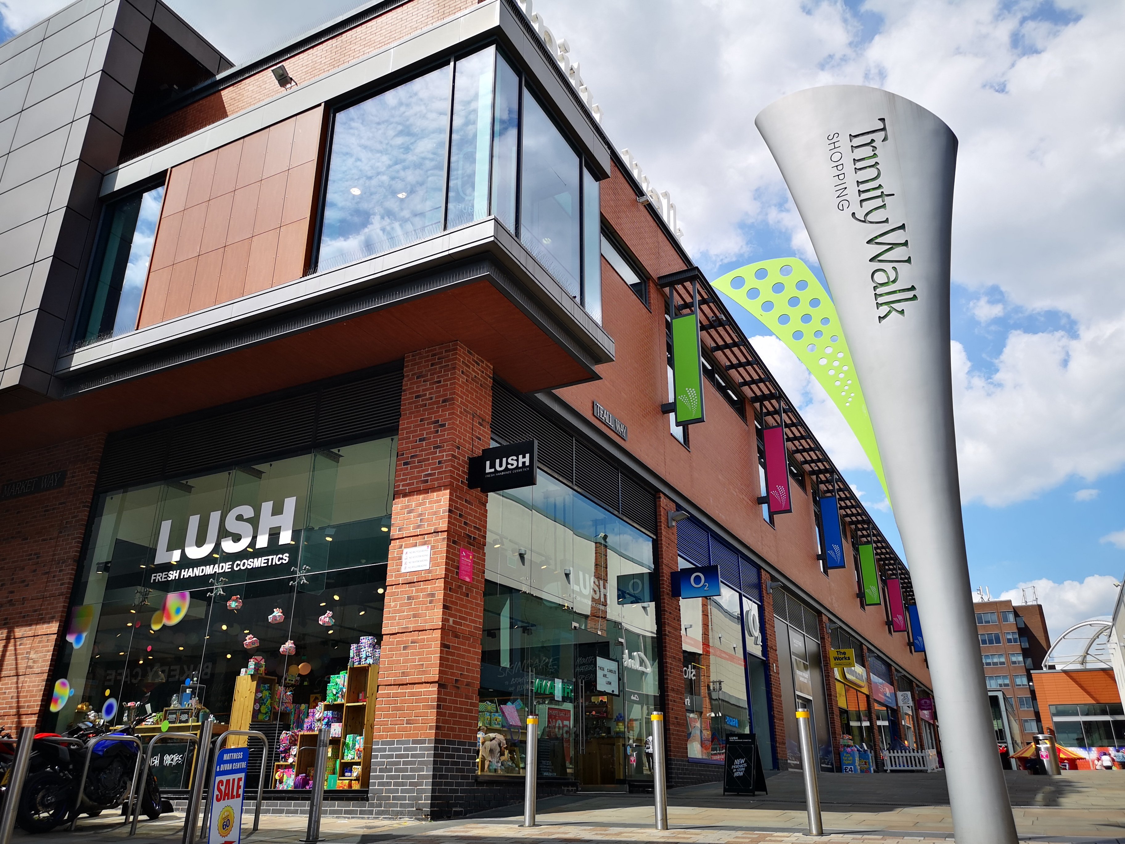 Exterior view of shop. Brick and glass frontage. Large entrance on left with illuminated Lush sign above. Products displayed in windows along full length of shop.