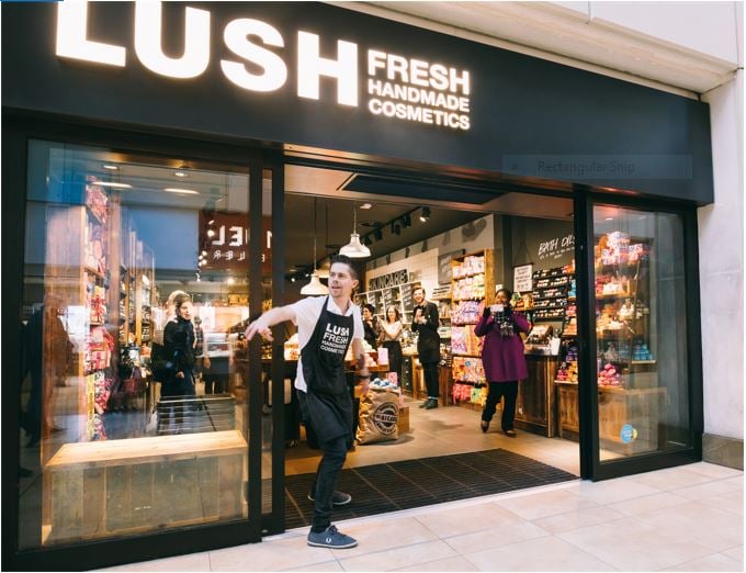 Exterior view of shop. Black framed shop with glass frontage. Large door way in centre with illuminated Lush sign above. Products displayed in windows either side of entrance. Member of staff stood in doorway.
