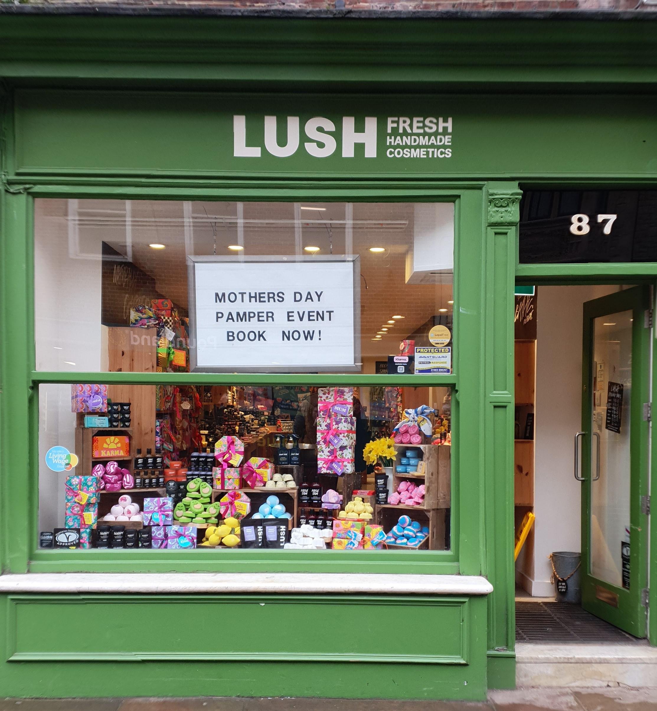 Exterior view of shop. Green framed shop with large glass window with Lush sign above. Door way on the right with a step up into the shop. Bright display of products in the window.