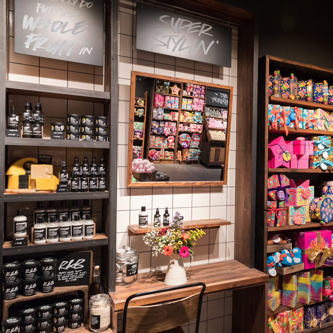 Haircare section on the shop-floor. Interior view of shop. Various packaged and unpackaged haircare products displayed on black shelves to the left next to a table with a chair and a large mirror. Signs above read We Really Do Put The Whole Fruit In, and Super Stylin'. To the right is a large display of varius brightly coloured giftsets.