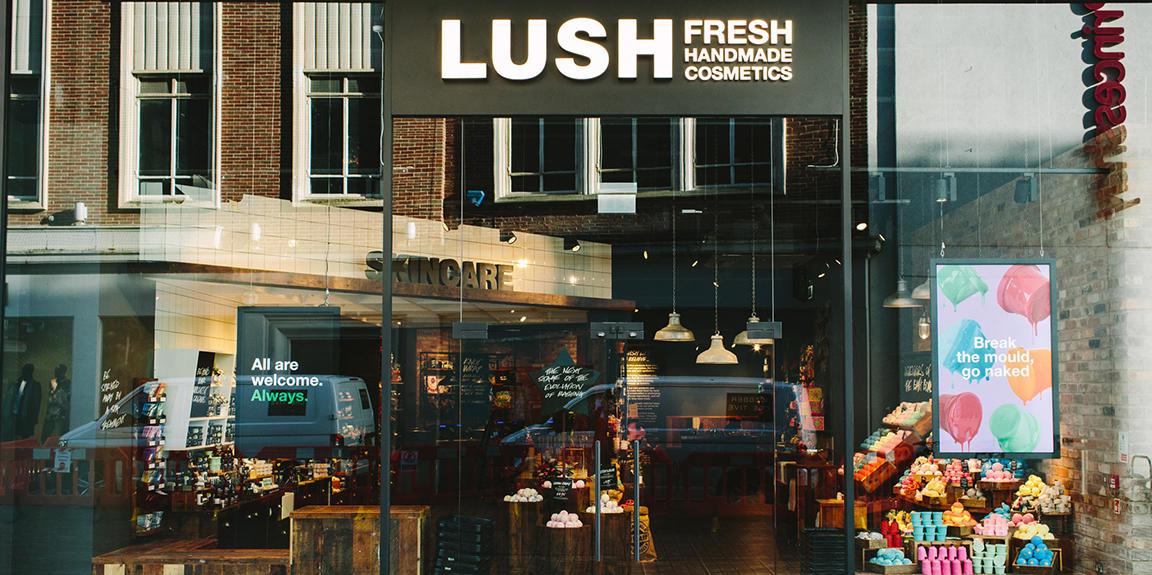 Exterior view of shop. Glass fronted shop with illuminated Lush sign above. Door way in centre with products displayed in windows either side of entrance.