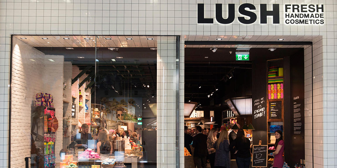 Manchester Arndale shop front. Exterior view of shop. White tiled shop with glass frontage. Large door way on right with illuminated Lush sign above. Products displayed in windows on left  side of entrance.