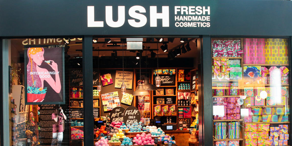 Warrington shop front. Exterior view of shop. Black framed shop with glass frontage. Large entrance in centre with illuminated Lush sign above. Products displayed in window to left of entrance. Abundant brightly coloured gift display in window to the right of the entrance.