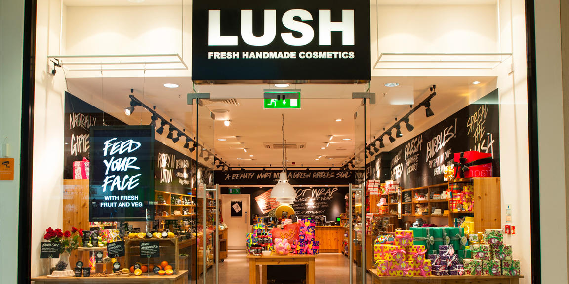 Exterior view of shop. Glass fronted shop. Large door way in centre with illuminated Lush sign above. Products displayed in windows either side of entrance.