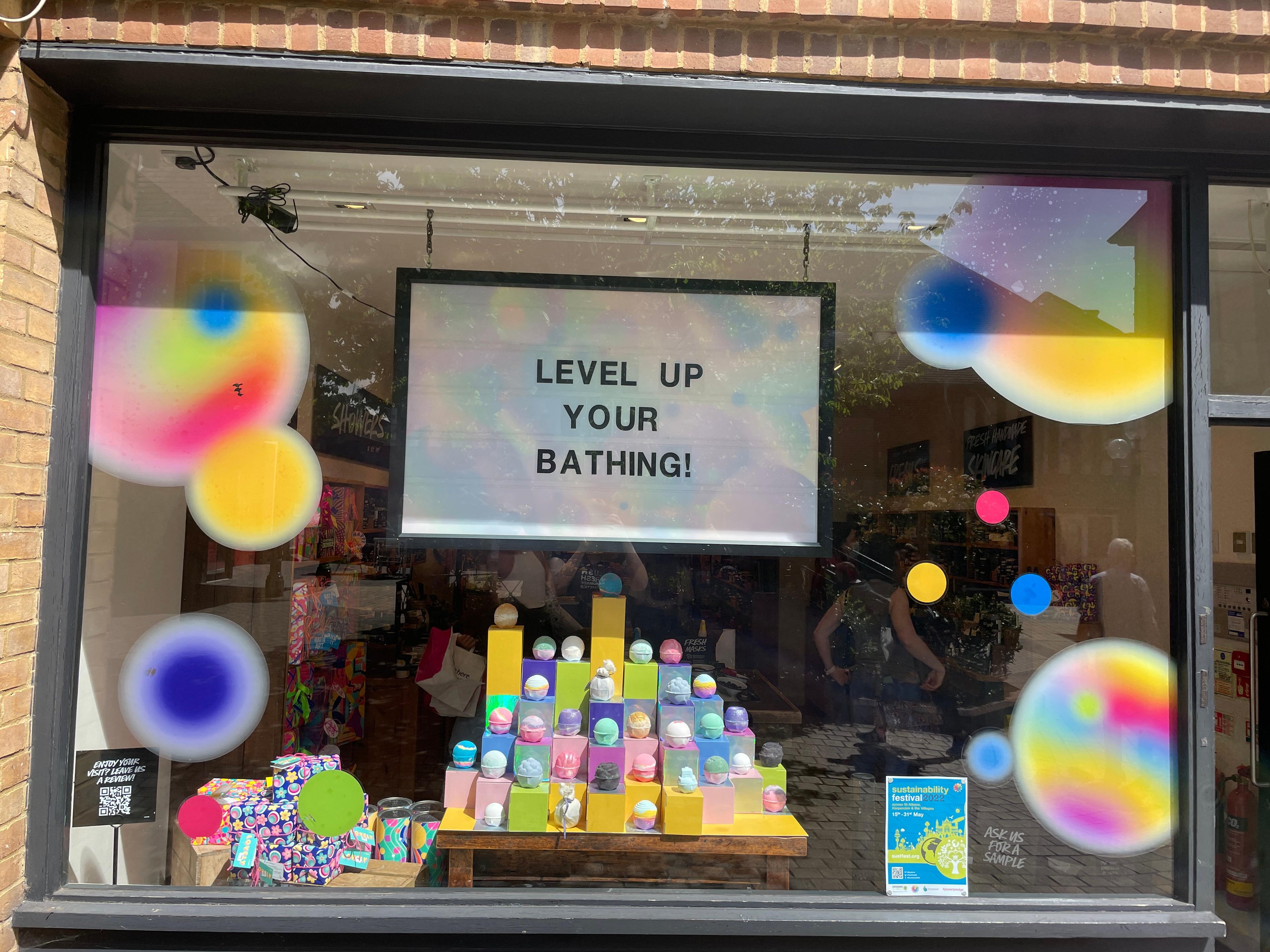 Exterior view of shop. Black framed shop 
 window. Products displayed in window below illuminated lightbox that reads Level Up Your Bathing.