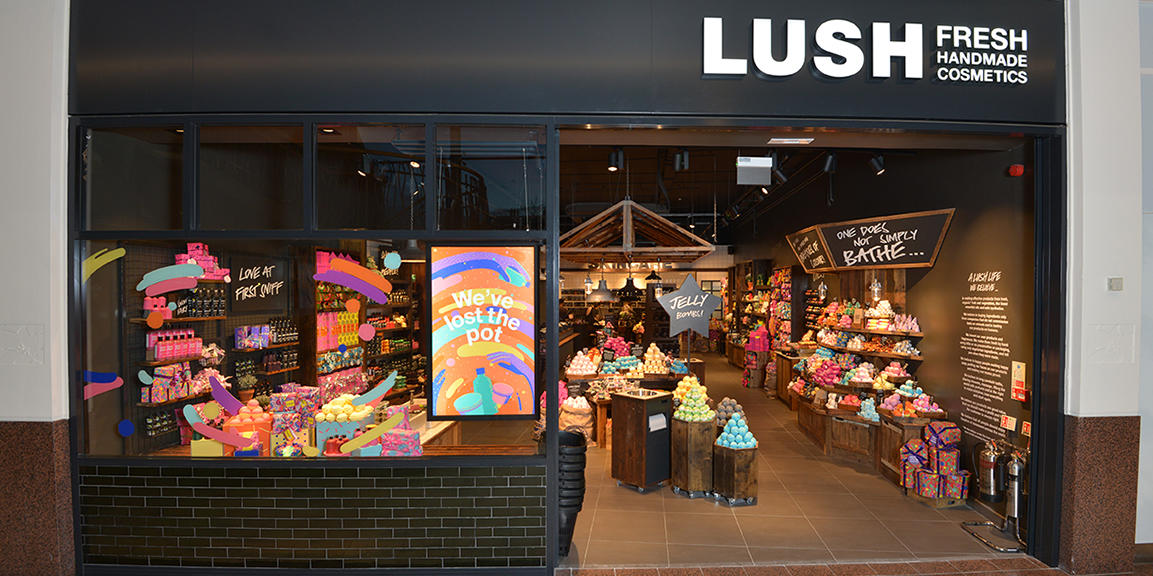 Exterior view of shop. Black framed shop with glass frontage. Large door way on right with illuminated Lush sign above. Products displayed in windows on the left of the entrance.