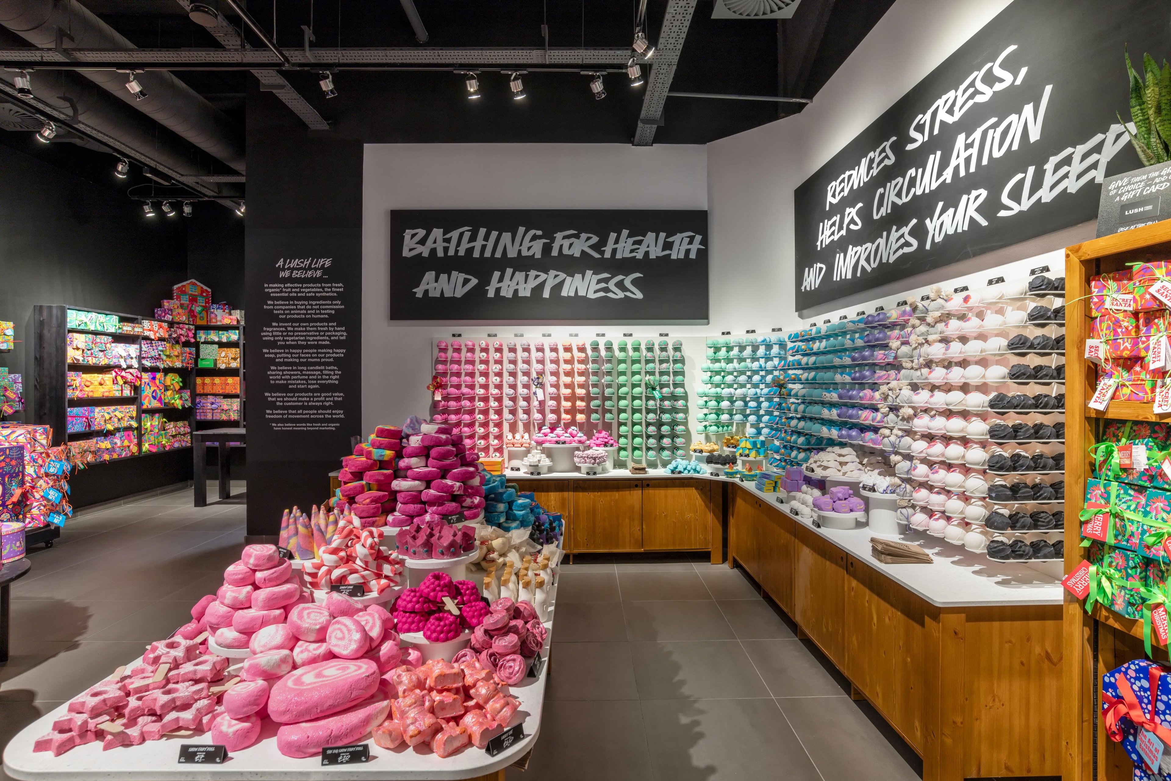 Interior view of shop. Central display island counting a pink themed display of various unpackaged bath products. On the walls on the right and behind are wall mounted displays of brightly coloured unpackaged bath products. Handwritten signs above.