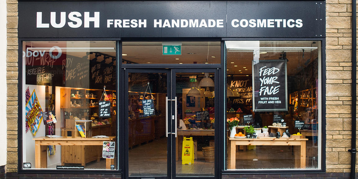 Exterior view of shop. Black framed shop with glass frontage. Large door way in centre with illuminated Lush sign above. Products displayed in windows either side of entrance.