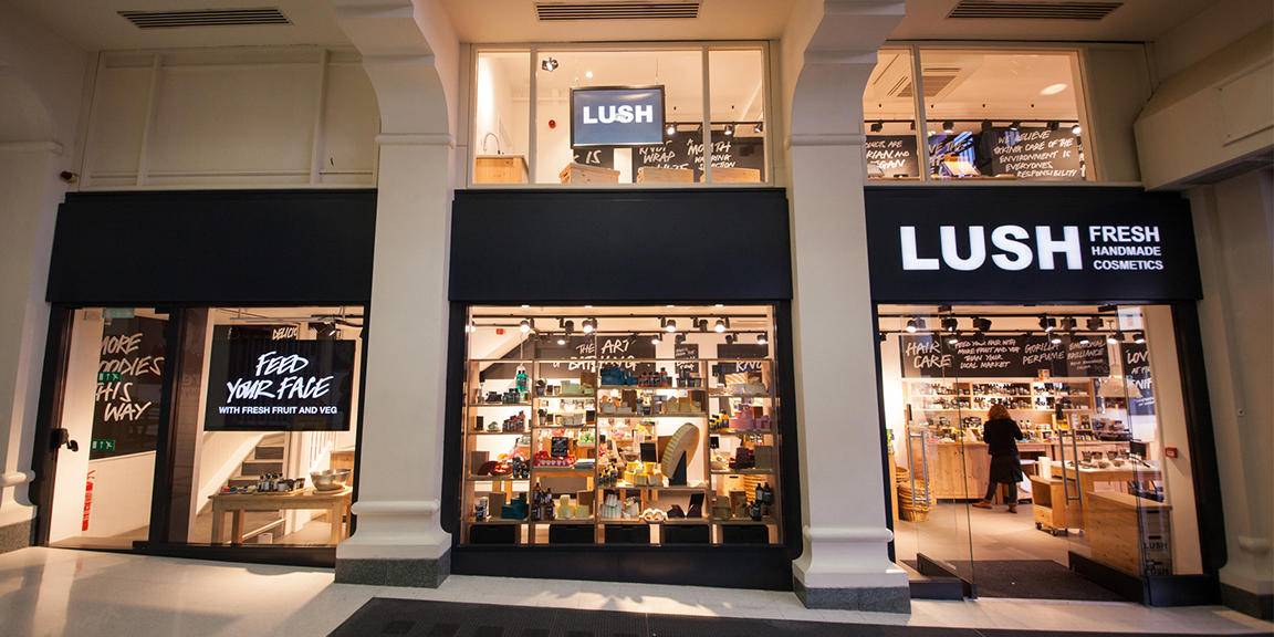 Tunbridge Wells shop front. Exterior view of shop. Black framed shop with glass frontage. Large entrance on the right with illuminated Lush sign above. Products displayed in windows to the left side of entrance.