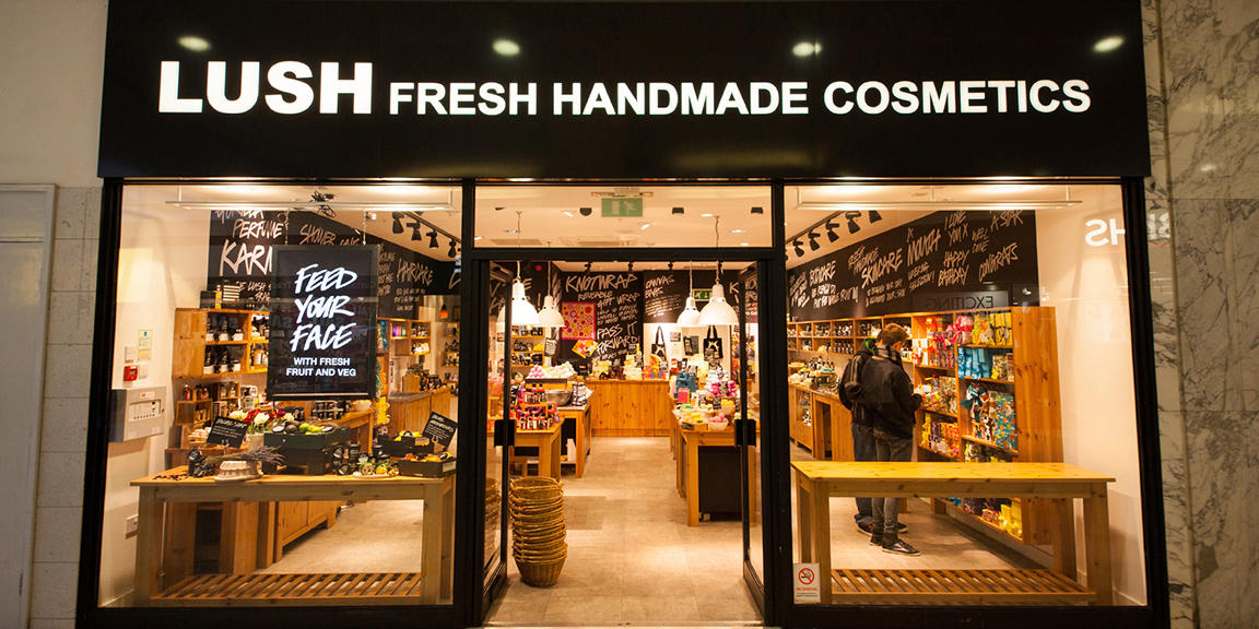 Exterior view of shop. Black framed shop with glass frontage. Door way in centre with illuminated Lush sign above. Products displayed in window on left side of entrance.