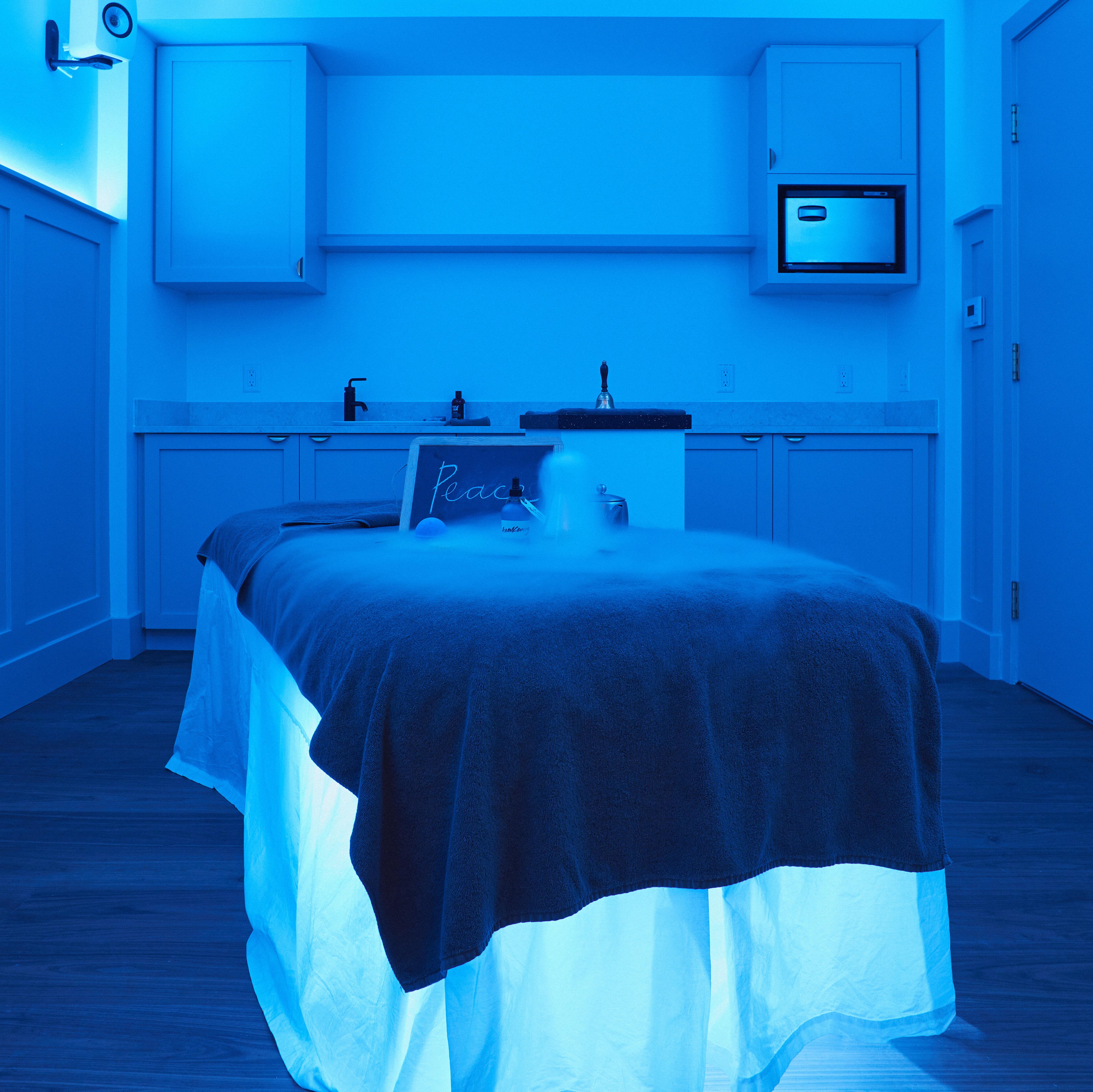 A treatment table is in the centre of a blue lit room. It has a dark blanket over a white sheet that drapes down to the floor. On the bed is a chalkboard with Peace handwritten on it. In front of the chalkboard are products wreathed in mist.
In the background are storage units with a sink.