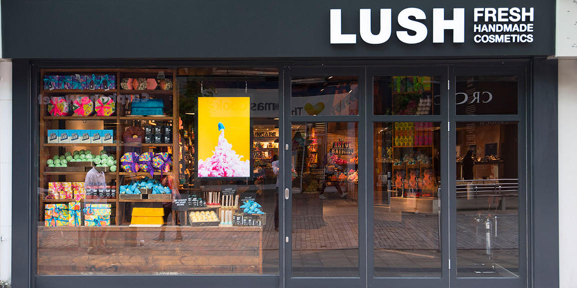 Swansea shop front. Exterior view of shop. Black framed shop with glass frontage. Large entrance on the right with illuminated Lush sign above. Products displayed in window to the left side of entrance.