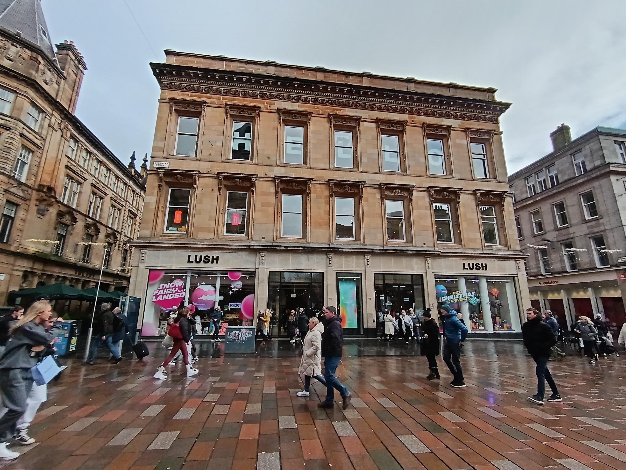 Exterior view of shop. Stand alone building on Buchanan Street. Yellow stone building with 3 storeys.  Large entrance in centre with  Lush sign above to the left. Products displayed in windows along length of shop front entrance. Large numbers of pedestrians passing by the front.