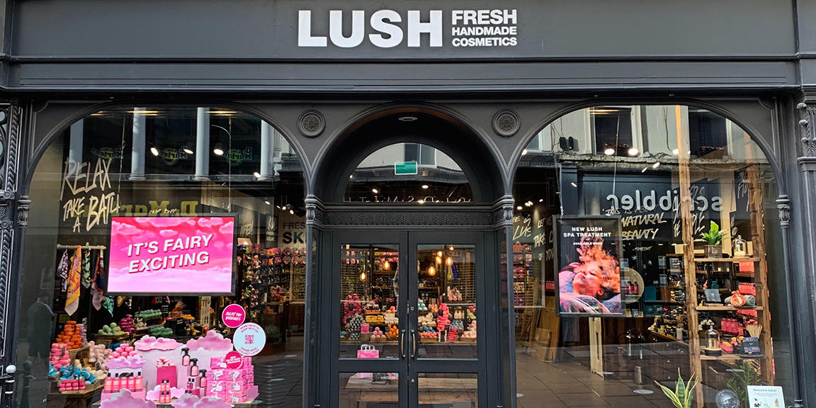 Bath shop front. Exterior view of shop. Black framed shop with glass frontage. Large entrance in centre with Lush sign above. Products displayed in windows either side of entrance.