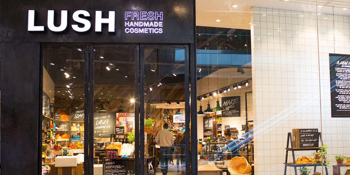 Stratford City shop front. Exterior view of shop. Black framed shop with glass frontage. Large entrance in centre with illuminated Lush sign above. Interior of shop is fully visible from outside of the shop.