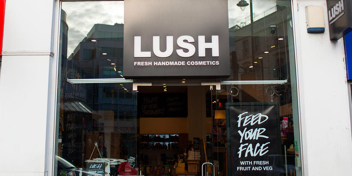 Wimbledon shop front. Exterior view of shop. Black framed shop with glass frontage. Large entrance in centre with illuminated Lush sign above. Sign in window to the right of the entrance reads Feed Your Face.