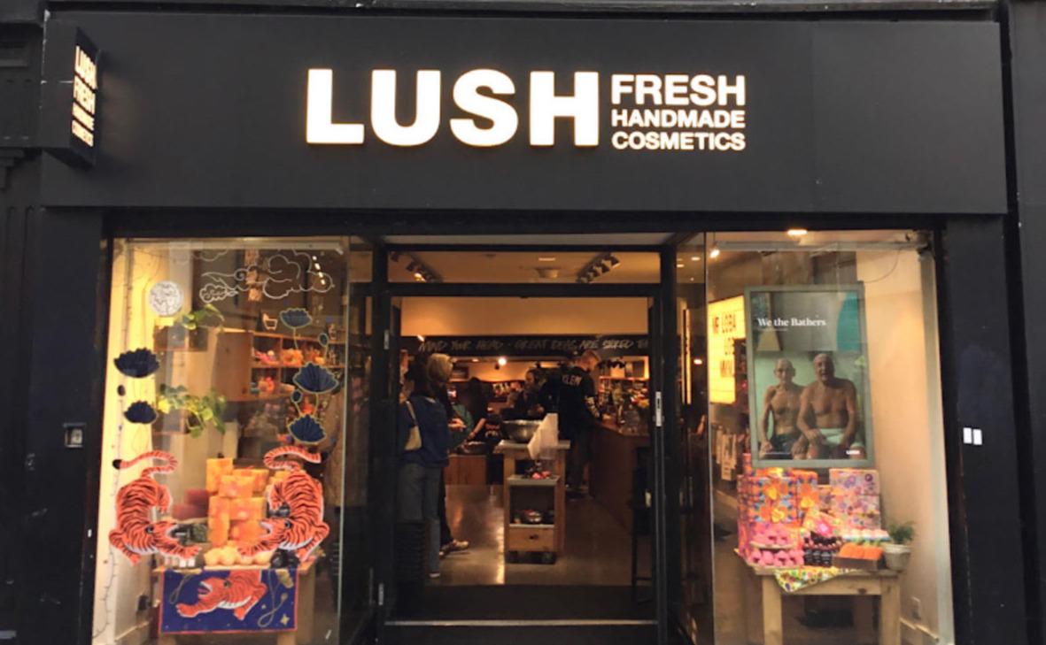 Exterior view of shop. Black framed shop with glass frontage. Large entrance in centre with illuminated Lush sign above. Products displayed in windows either side of entrance.