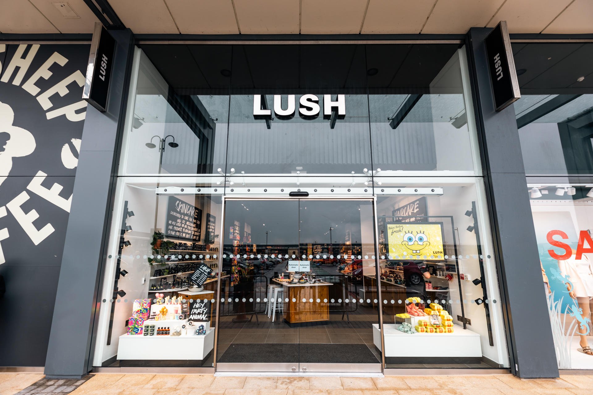 Exterior view of shop. Black framed shop with glass frontage. Large door way in centre with illuminated Lush sign above. Products displayed in windows either side of entrance.