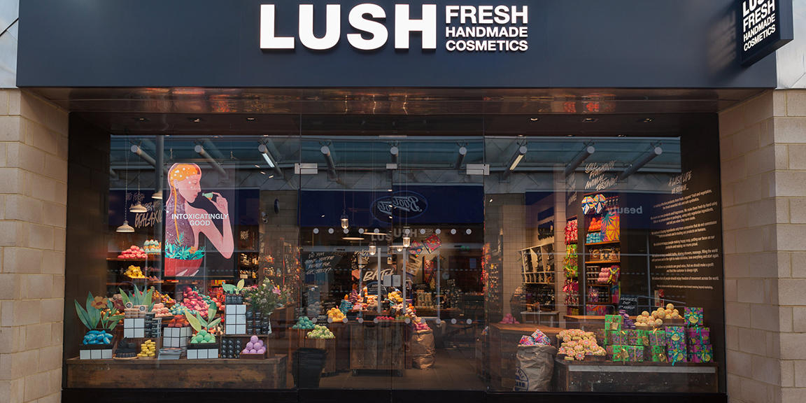 Exterior view of shop. Black framed shop with glass frontage. Large door way in centre with illuminated Lush sign above. Products displayed in windows either side of entrance.