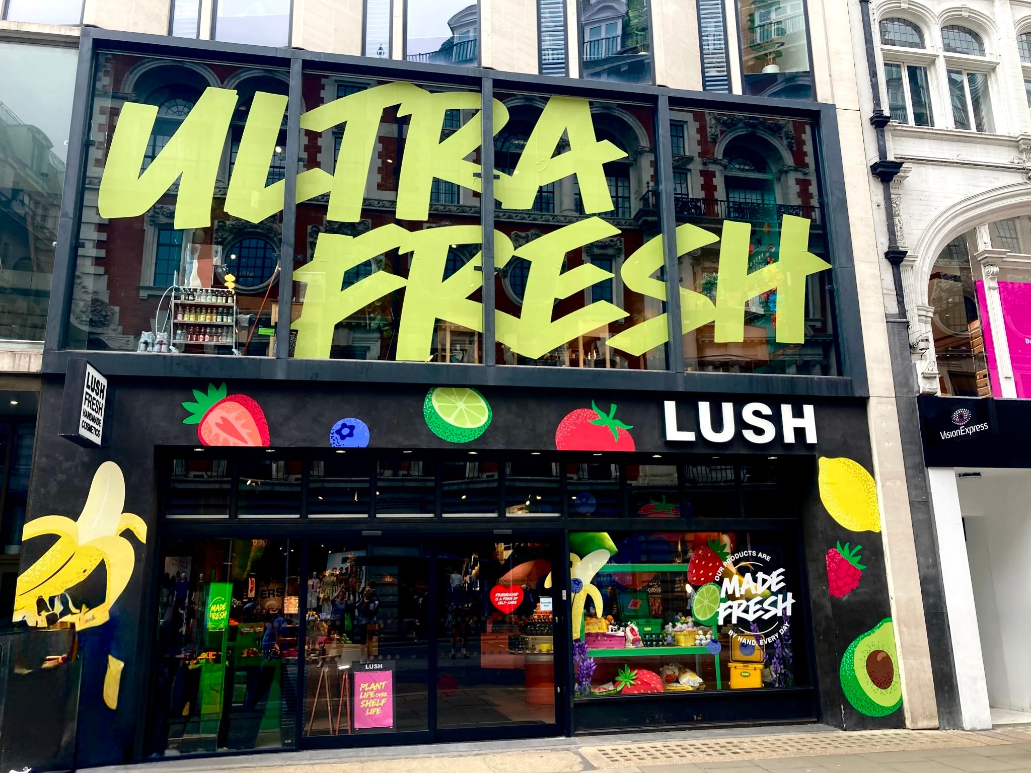 Lush Oxford Street shop front. Exterior view of shop. Black framed shop with glass frontage. Large entrance on left side with illuminated Lush sign above on the right. Products displayed in window to the right side of entrance.