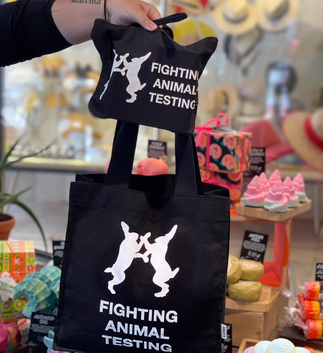 A black cotton makeup bag and a black cotton tote bag both displaying the fighting animal testing logo and the words Fighting Animal Testing are held up in front of a display of packaged and unpackaged products.