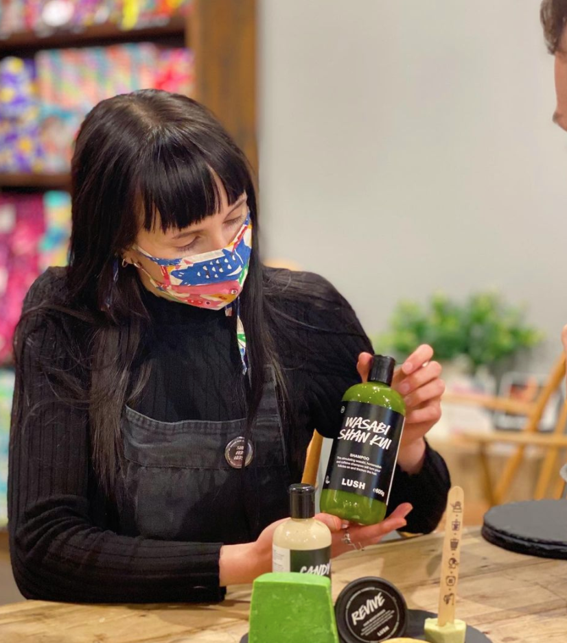 Member of staff wearing a face mask, holding a bottle of Wasabi Shan Kui shampoo towards the camera. Various packaged and unpackaged haircare products in front of her.