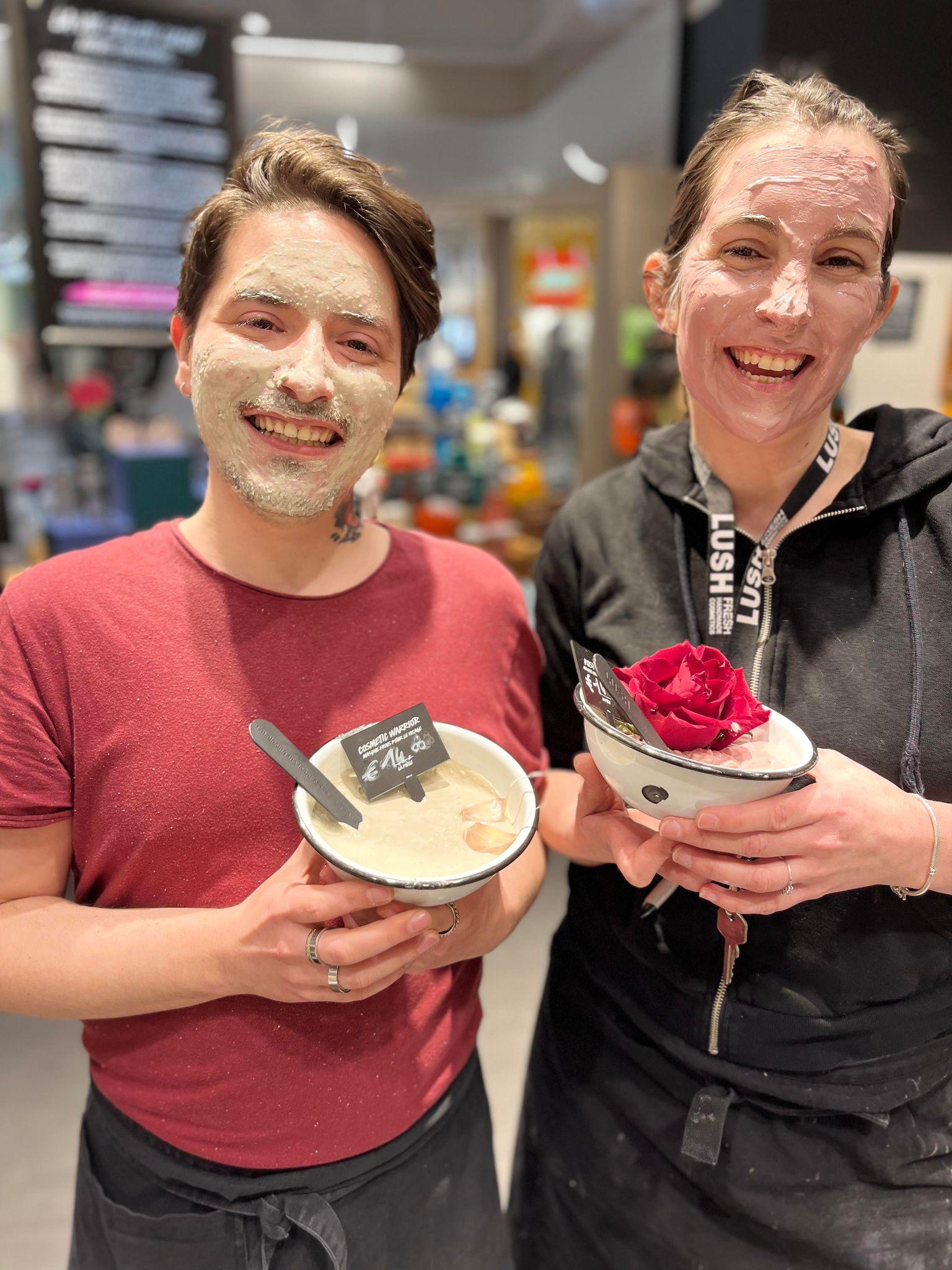 Two members of staff smiling. Both have fresh facemasks covering their faces, and they are each holding a bowl of facemask product.