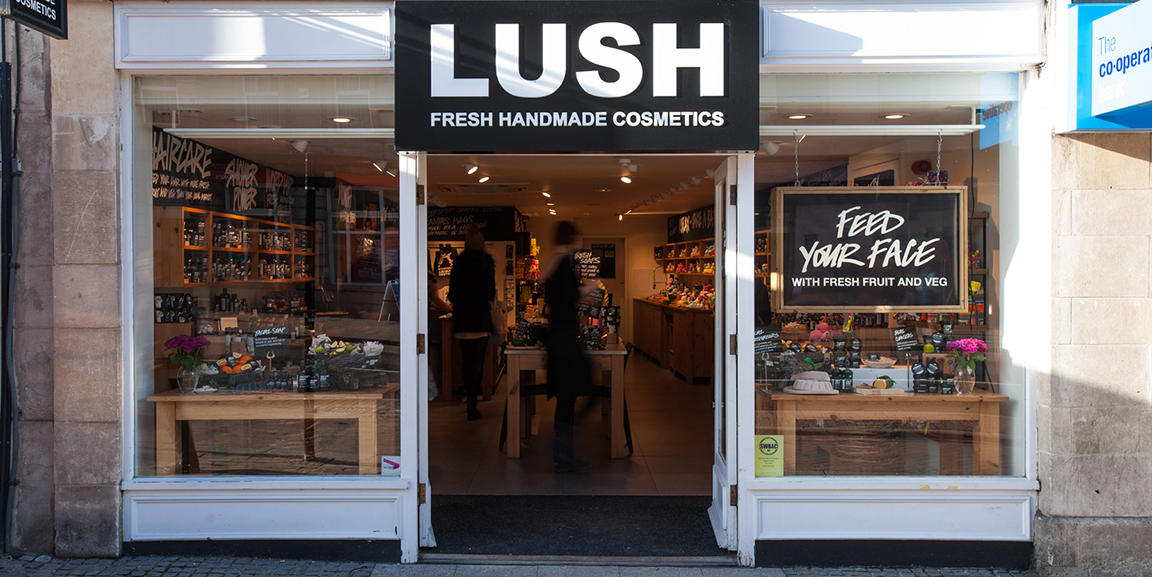 Taunton shop front. Exterior view of shop. White framed shop with glass frontage. Large entrance in centre with Lush sign above. Products displayed in windows either side of entrance. Sign in right side window reads Feed Your Face.