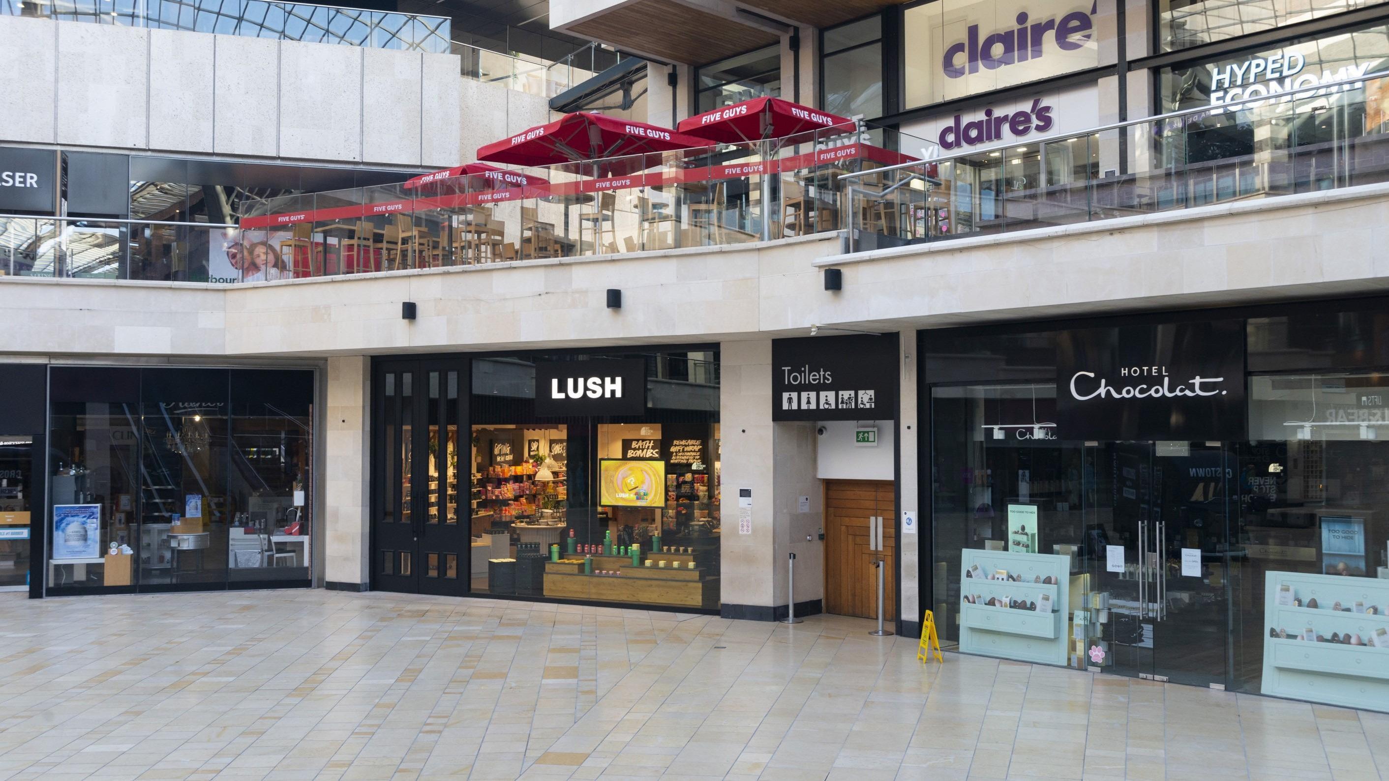 Exterior view of shop showing location within shopping centre. Black framed glass front with illuminated Lush sign above entrance.