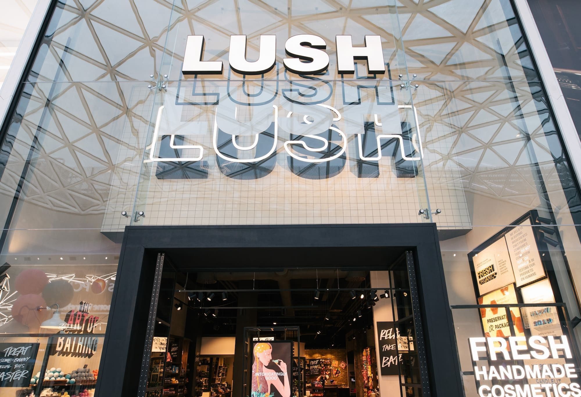 Exterior view of shop. Glass fronted shop. Large entrance in centre with large illuminated Lush sign above. Full interior of the shop can be seen from the outside.