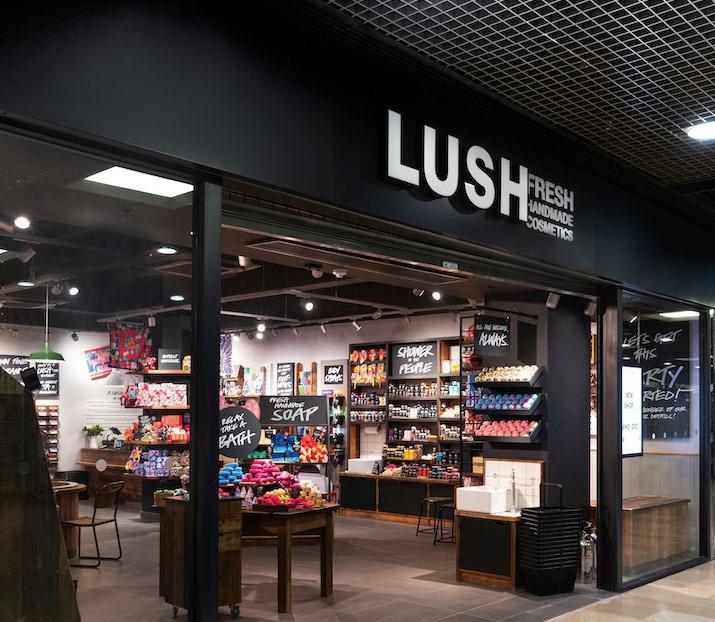 Peterborough Store Front. Exterior view of shop. Black framed shop with glass frontage. Large entrance in centre with illuminated Lush sign above. Full interior of the shop is visible from the outside.