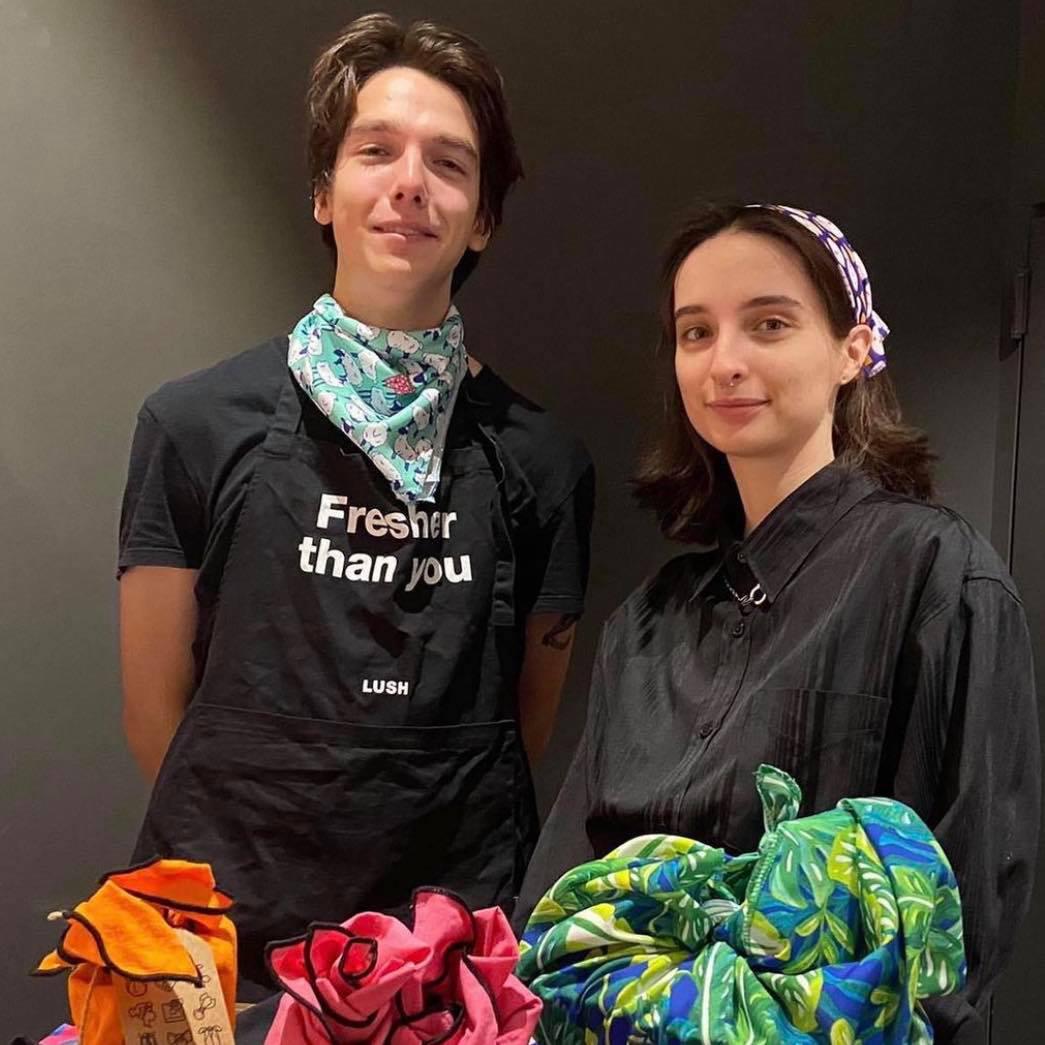 2 members of staff dressed in black are stood in front of gifts wrapped with Knotwrap scarves. Both are also wearing knot wraps.