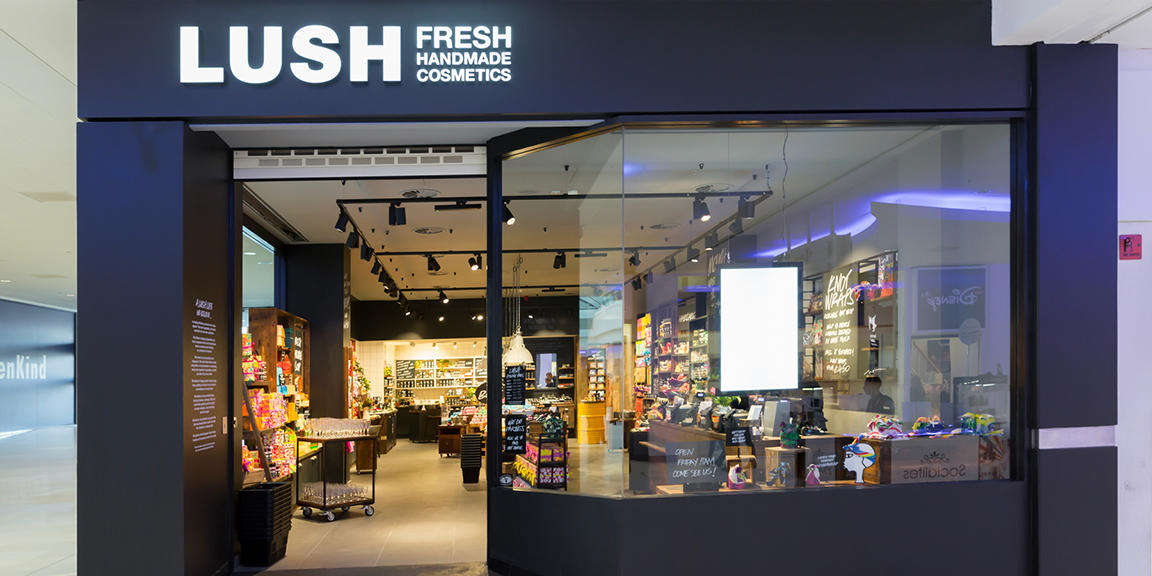 Lush Portsmouth's Store Front in Cascades Shopping Centre. Exterior view of shop. Black framed shop with glass frontage. Large entrance on left with illuminated Lush sign above. Products displayed in window to right side of entrance.